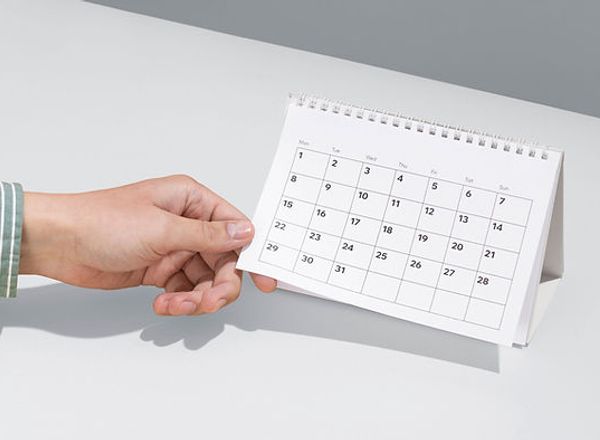 Hand zeigt auf einen weißen Tischkalender auf hellem Schreibtisch, Terminplanung wird symbolisiert