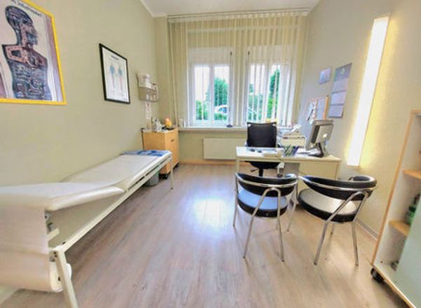 Heller Behandlungsraum mit weißer Untersuchungsliege, Schreibtisch mit Computer und zwei schwarzen Stühlen vor großem Fenster