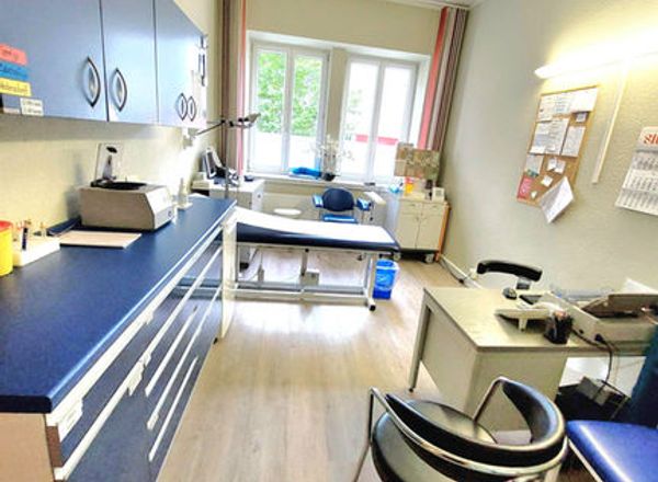 Moderner Behandlungsraum einer Arztpraxis mit blauer Einrichtung, Behandlungsliege und großem Fenster