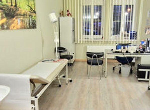 Heller Behandlungsraum mit weißer Liege, Schreibtisch und großen Fenstern mit Lamellenvorhängen